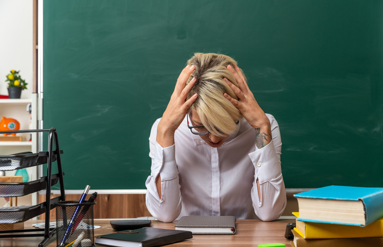 Stressed Young Blonde Female Teacher Wearing Glasses Sitting Desk With School Tools Classroom Keeping Hands Head